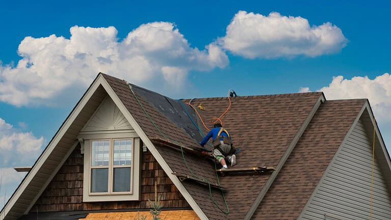 A person in blue working on a house roof, brown shingles, blue sky with white clouds in background