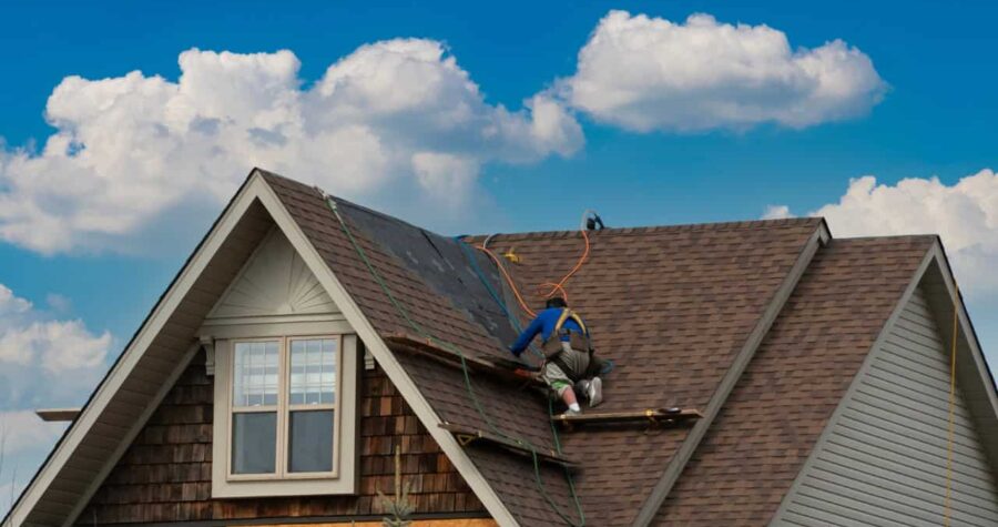 A person in blue working on a house roof, brown shingles, blue sky with white clouds in background