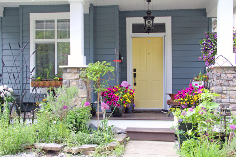Front porch with vibrant flower pots, blooming petunias and zinnias, arranged on steps and by the doorway, blue house exterior, yellow front door, stone column accents, white trim, hanging lantern