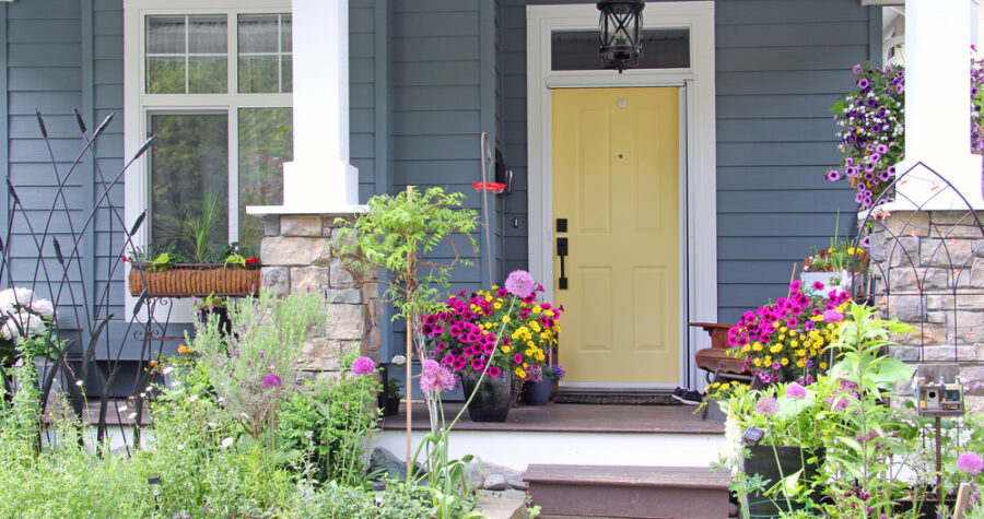 Front porch with vibrant flower pots, blooming petunias and zinnias, arranged on steps and by the doorway, blue house exterior, yellow front door, stone column accents, white trim, hanging lantern