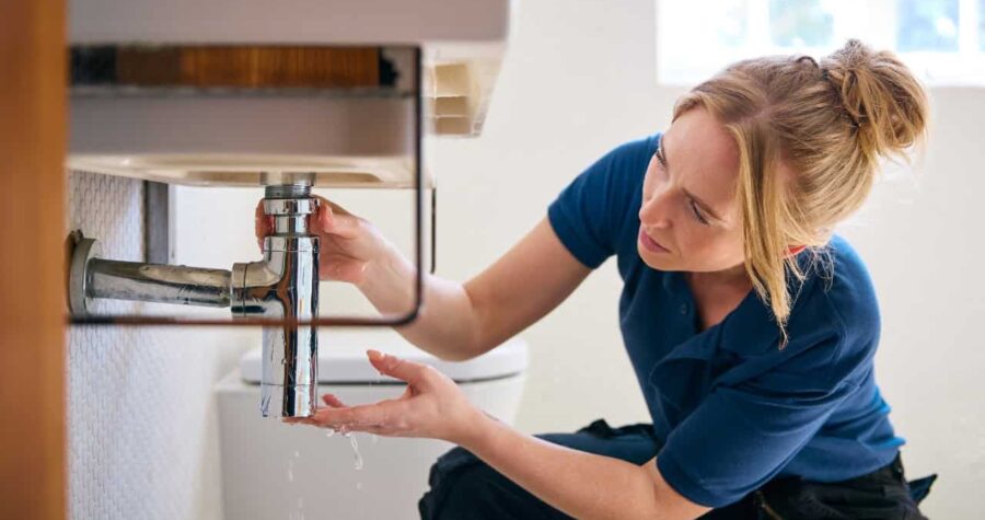 Person in blue shirt crouching near bathroom sink, examining or repairing faucet plumbing, hands positioned under sink fixture, bathroom interior with mirror visible in background