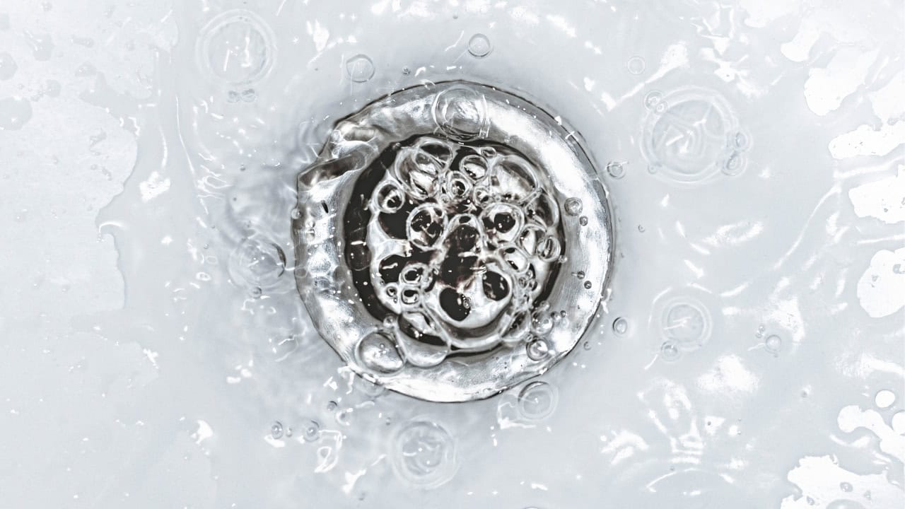 Close-up of a silver sink drain with water swirling and bubbling around it, light reflecting off the metal