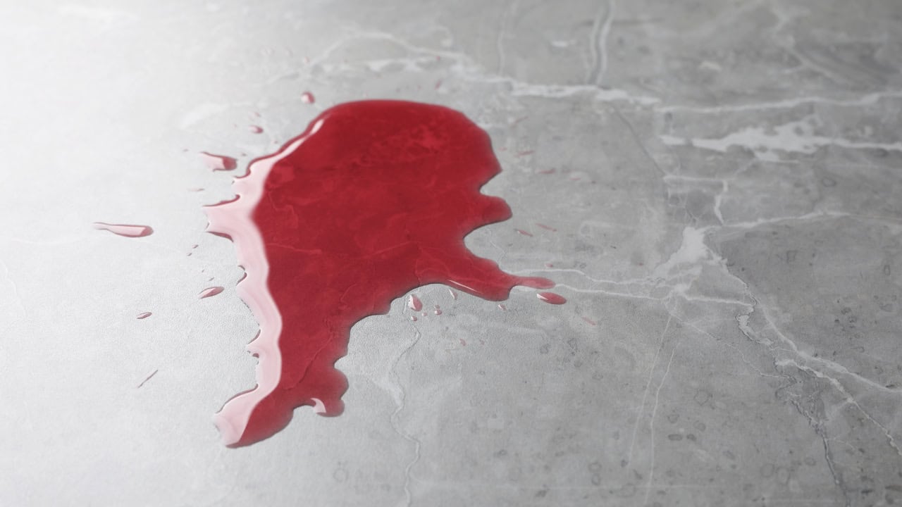 A spilled red liquid on a shiny gray surface, liquid pooling and spreading, reflecting light on the smooth surface, contrasting color of the red liquid against the neutral background