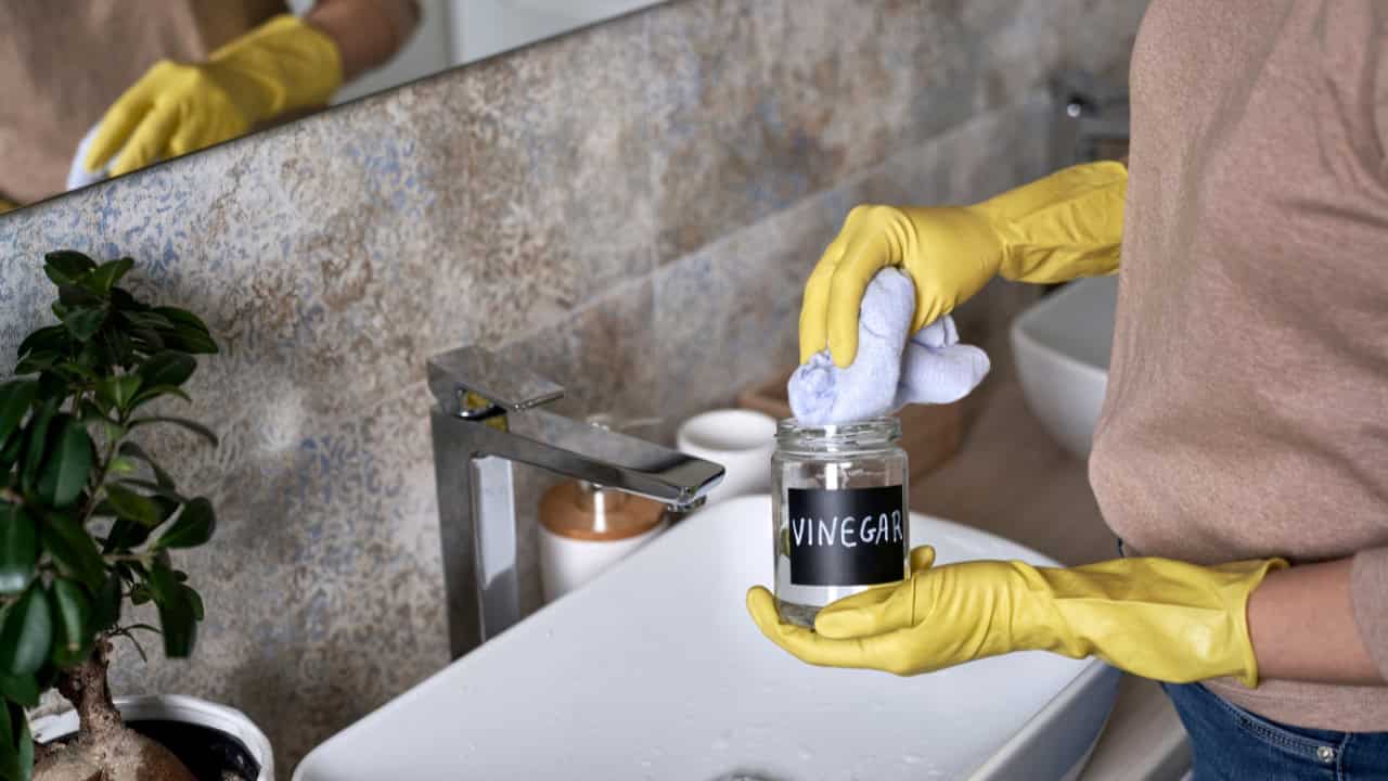 Person wearing yellow gloves cleaning a bathroom sink with a cloth, holding a jar labeled "vinegar," using natural cleaning methods