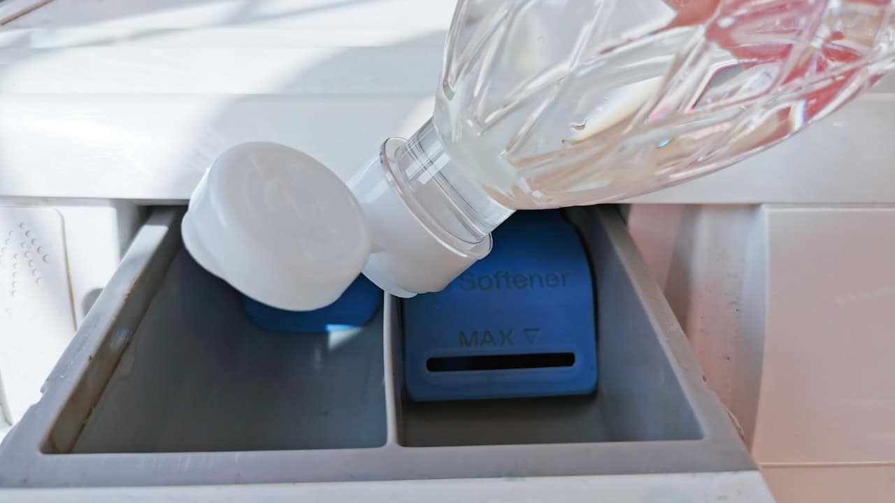 Person pouring liquid from a plastic bottle into a washing machine detergent dispenser drawer, blue appliance visible, close-up view
