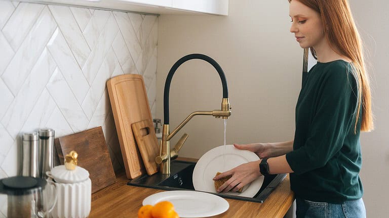 User washing dishes, hands holding a plate under running water, kitchen sink, dishwashing process, cleaning plate with sponge, modern kitchen setup, wooden countertop