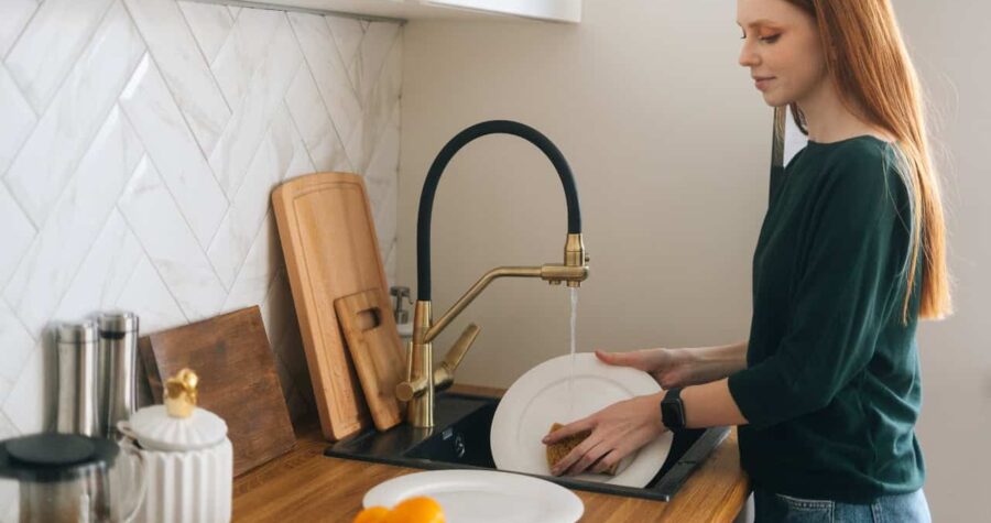 User washing dishes, hands holding a plate under running water, kitchen sink, dishwashing process, cleaning plate with sponge, modern kitchen setup, wooden countertop