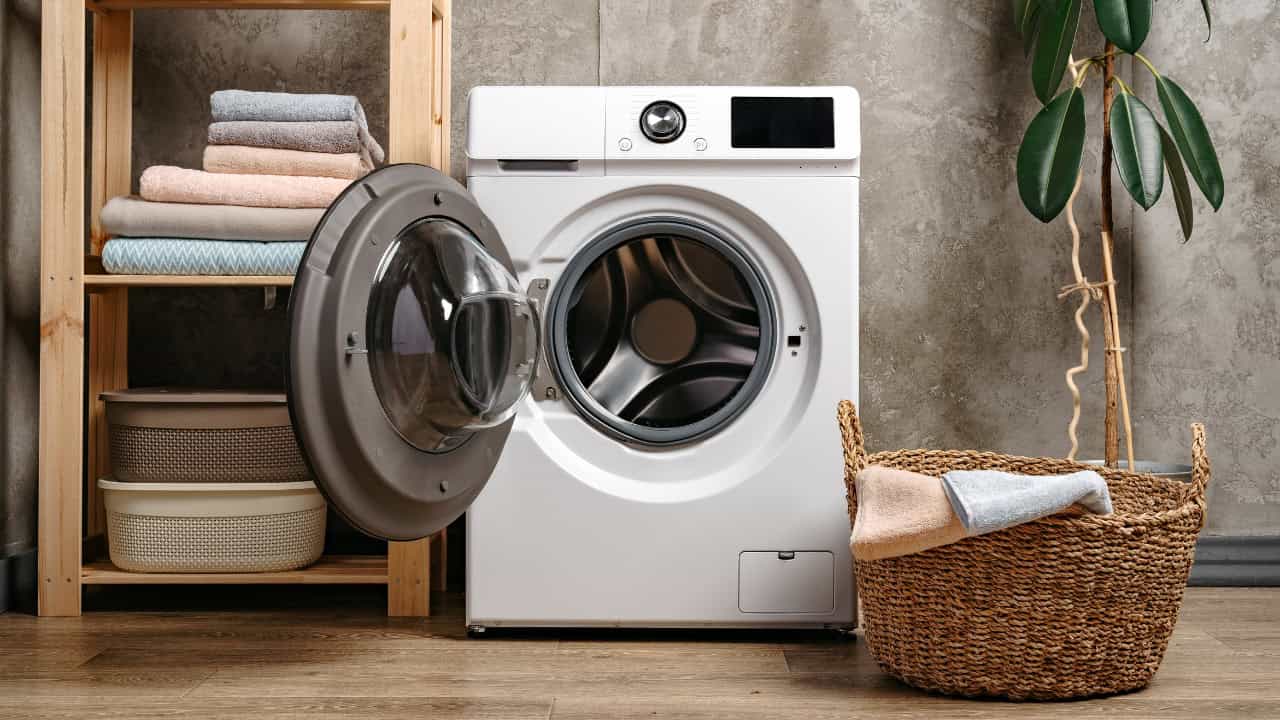Front-loading washing machine, open door, modern laundry room, shelves with neatly folded towels, wicker basket with clothes, concrete-textured wall, digital control panel, clean and organized space