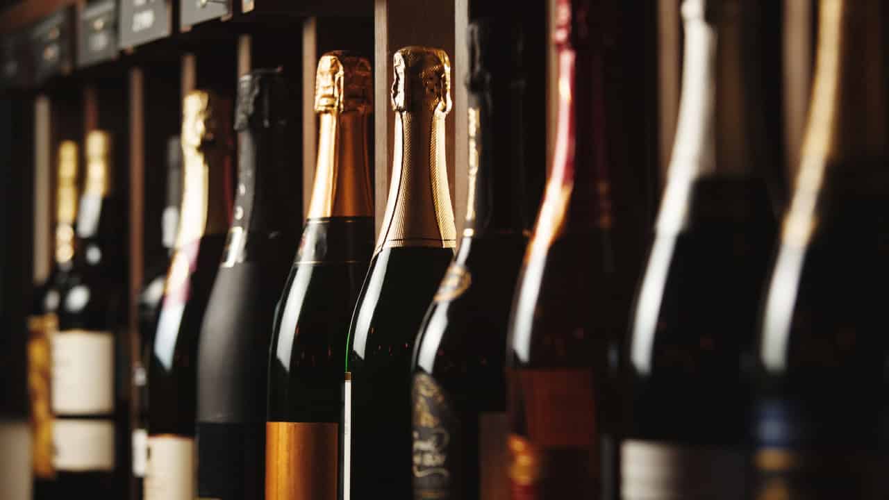 A row of champagne and wine bottles, gold and black foil tops, neatly arranged on a shelf, dim warm lighting