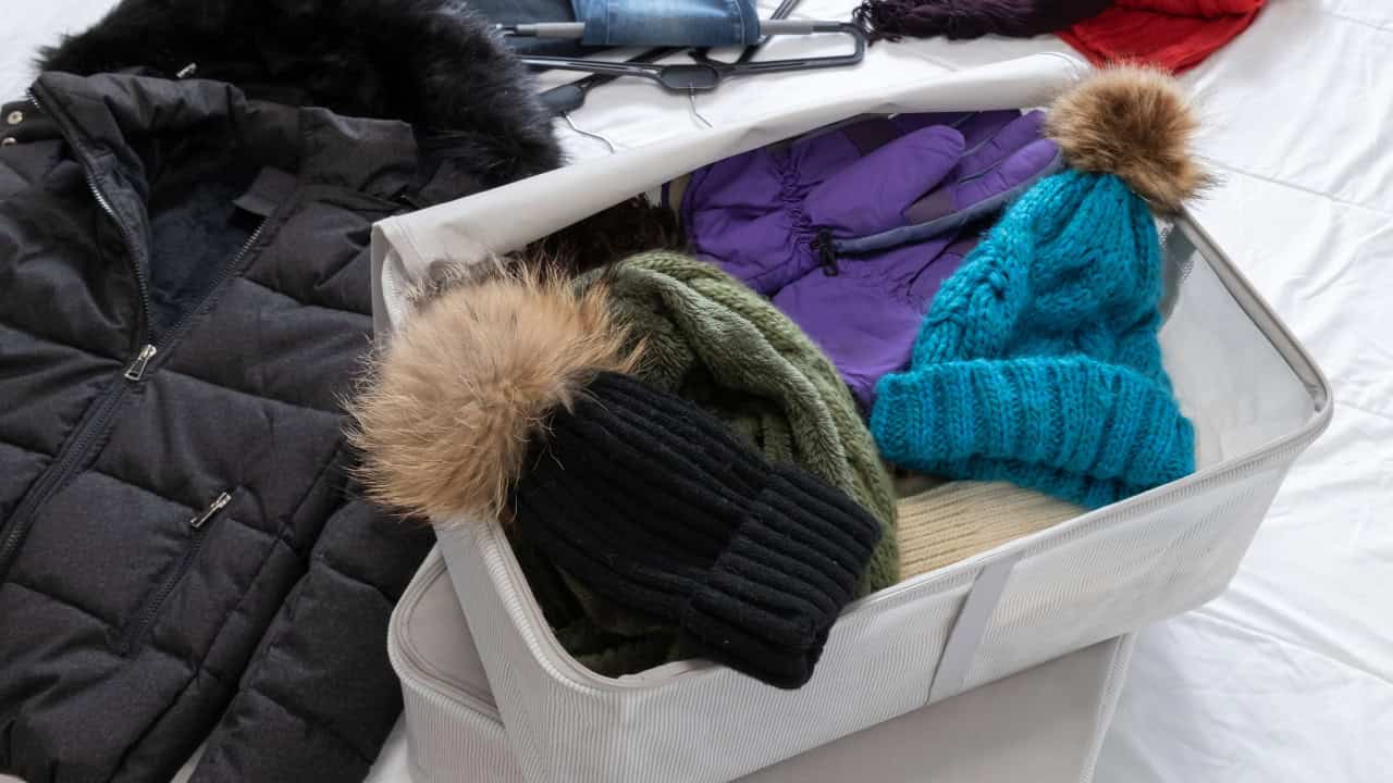 A storage box filled with winter accessories, a black knit hat with a fur pom-pom, green and purple scarves, purple gloves, and a blue knitted sweater visible, neatly packed cold weather clothing