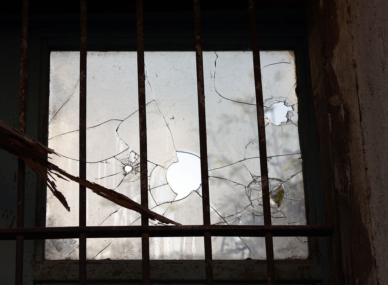 The image shows a broken window with shattered glass, some pieces have holes, and the window is behind a rusty metal grate, parts of the window frame are damaged, the glass has cracks, the surrounding area is slightly worn, the window appears to be old and neglected