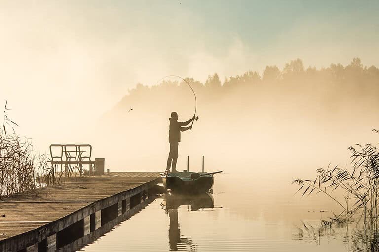 A man fishing alone, standing on a small boat near a wooden dock, casting his fishing line into a calm lake, surrounded by mist, with trees faintly visible in the background, early morning atmosphere