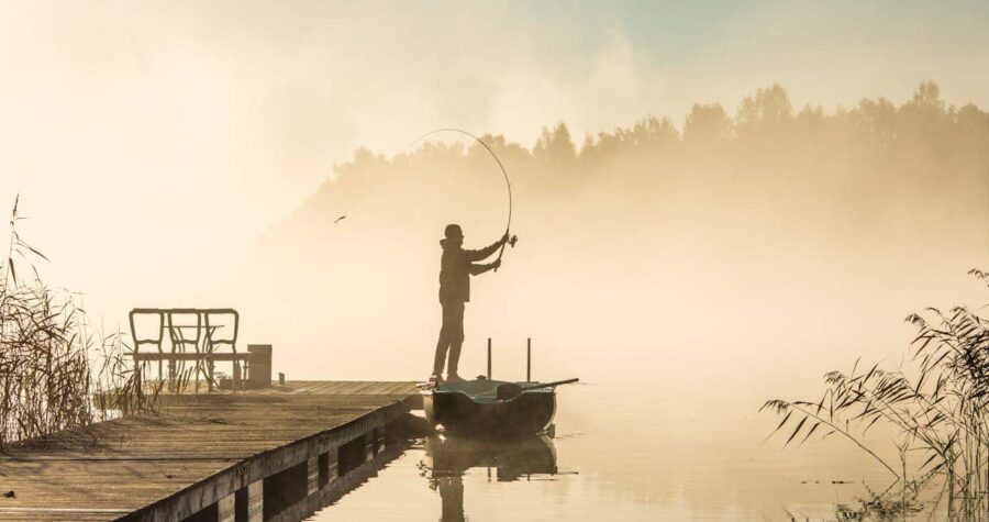 A man fishing alone, standing on a small boat near a wooden dock, casting his fishing line into a calm lake, surrounded by mist, with trees faintly visible in the background, early morning atmosphere