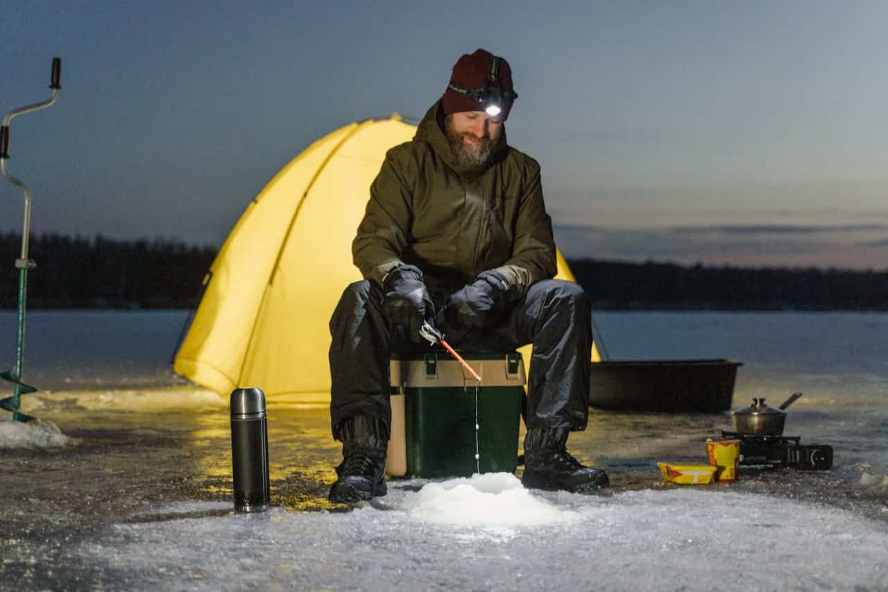 A man ice fishing at night, wearing a winter jacket, gloves, and a headlamp, sitting on a cooler, holding a fishing rod over a hole in the ice, a yellow tent glowing behind him, a thermos on the ice, cooking equipment and food nearby