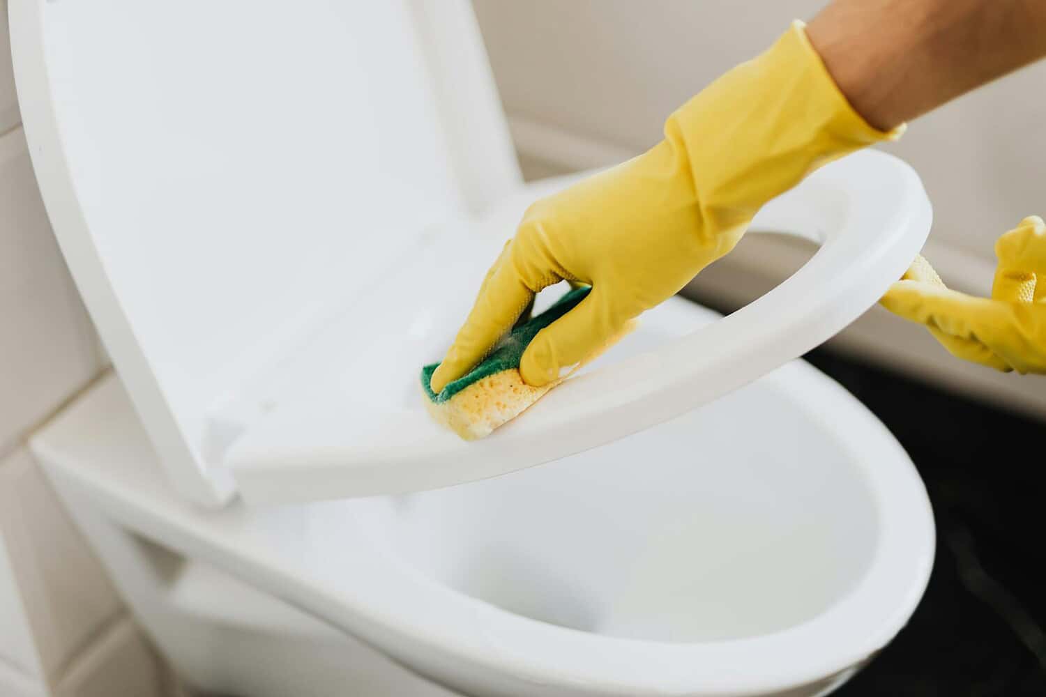 A person using yellow gloves and a sponge to clean a toilet seat indoors.