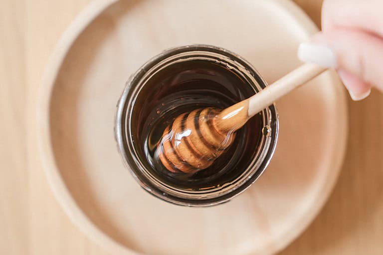 Glass jar filled with dark honey, wooden honey dipper partially submerged, placed on a beige surface