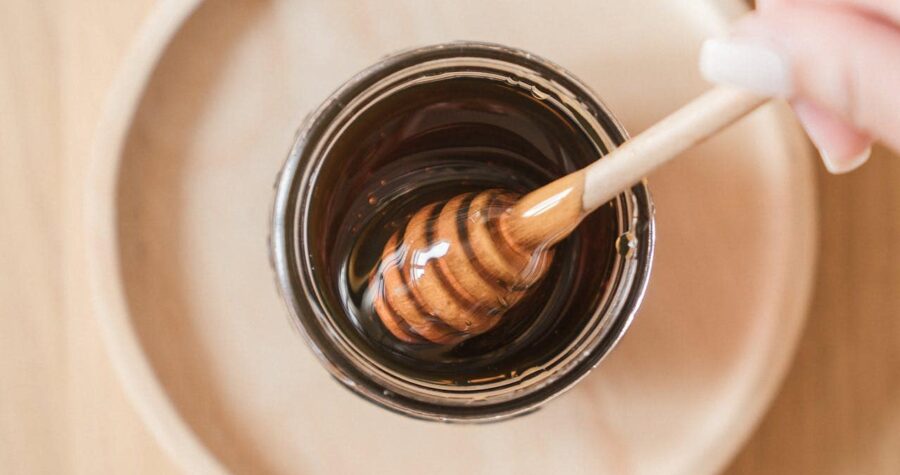 Glass jar filled with dark honey, wooden honey dipper partially submerged, placed on a beige surface