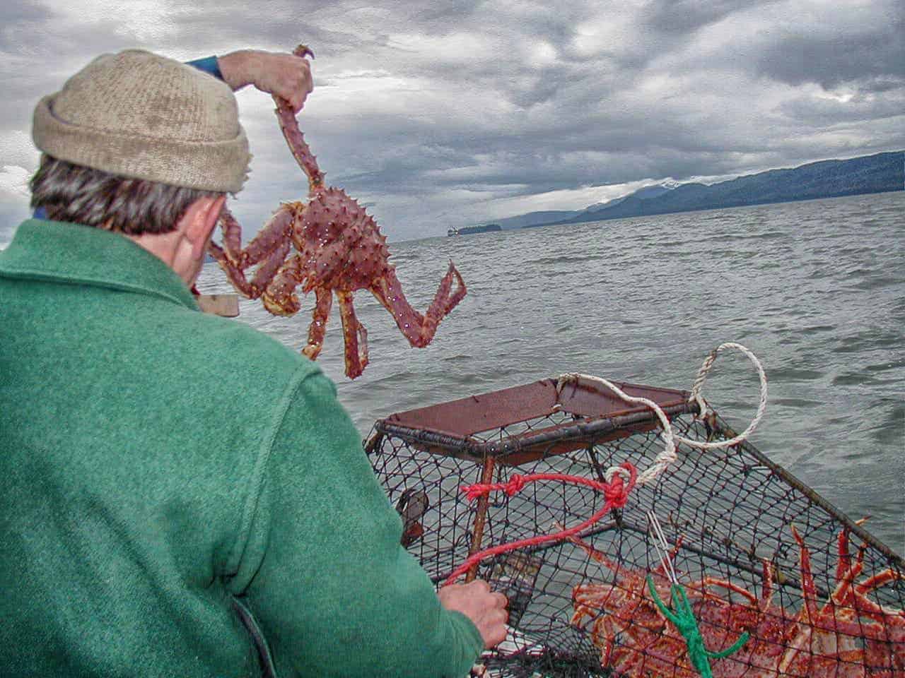A person in a green jacket and knit hat holding up a large red king crab while on a fishing boat, with crab traps visible and a gray cloudy sky over water with distant mountains in the background