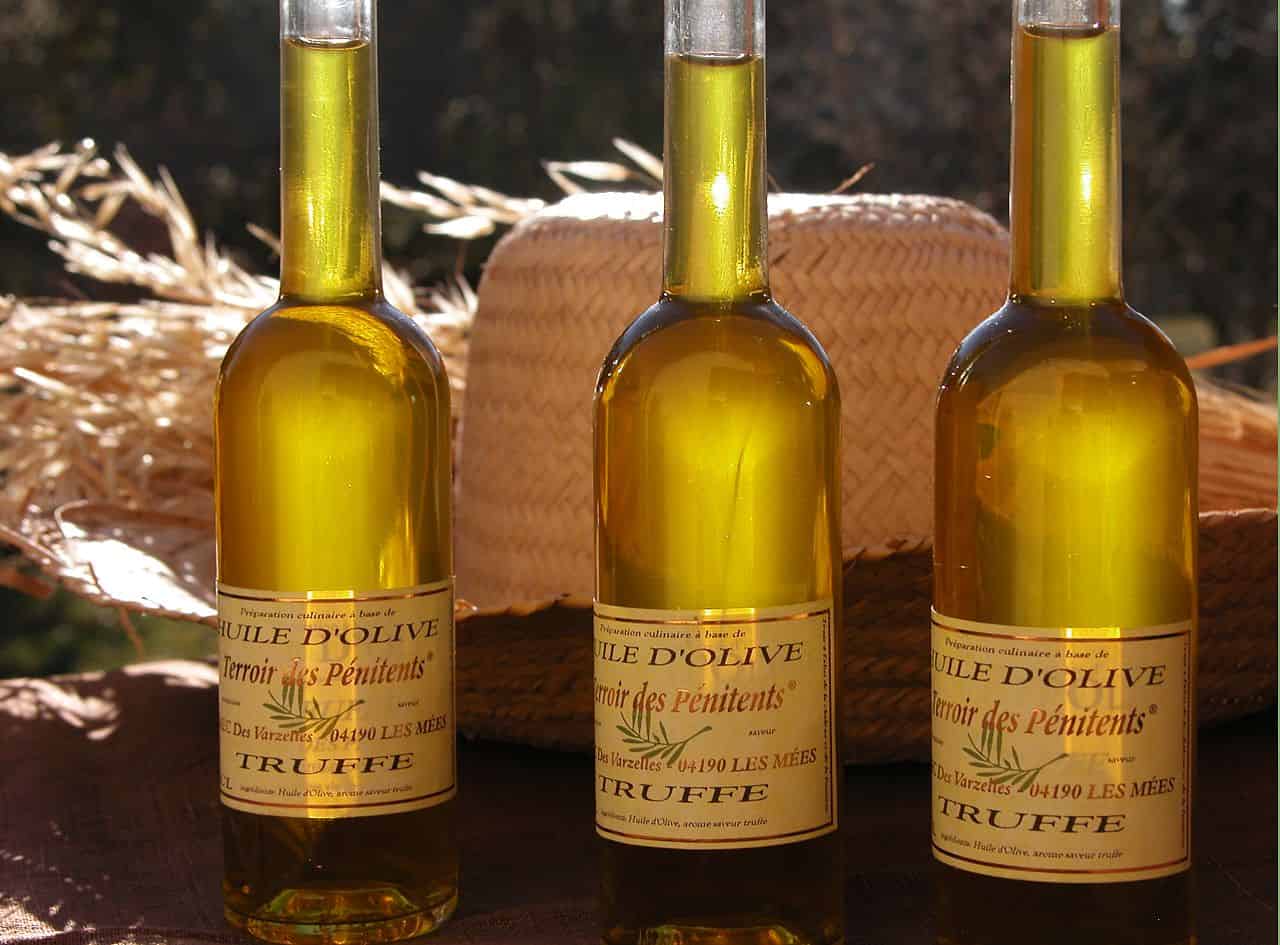 Three bottles of truffle-infused olive oil are arranged on a table with a straw hat in the background. The labels on the bottles read "Huile d'Olive Terroir des Penitents" and include details about the product’s origin and ingredients