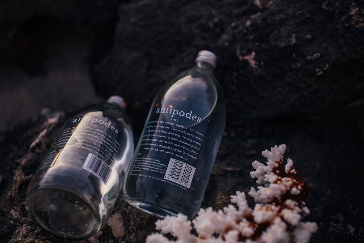 Two empty Antipodes water bottles lying on dark rocks, soft lighting, reflective glass, coral fragment nearby, outdoor setting, eco-friendly packaging