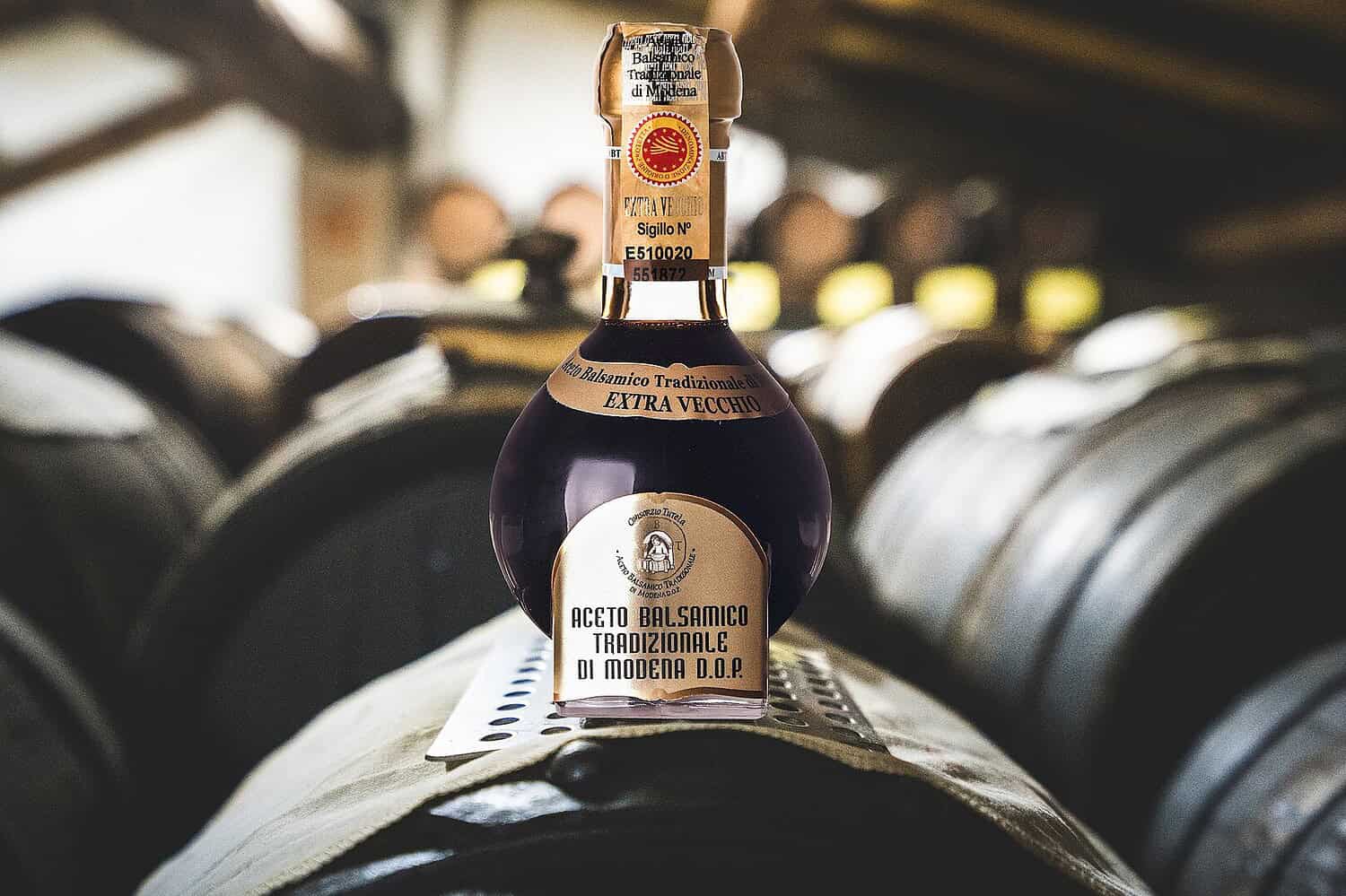 Bottle of traditional balsamic vinegar from Modena, labeled "Extra Vecchio," placed on a wooden barrel, aged barrels in the blurred background, warm lighting, artisanal production setting