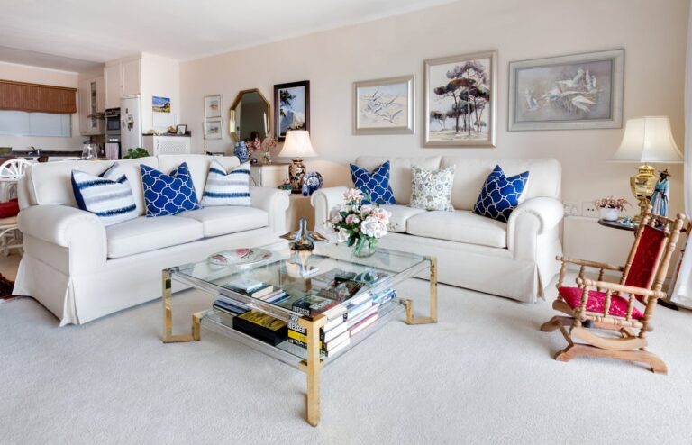 A well-decorated living room featuring two white sofas with blue patterned cushions, a glass coffee table with books, floral arrangements