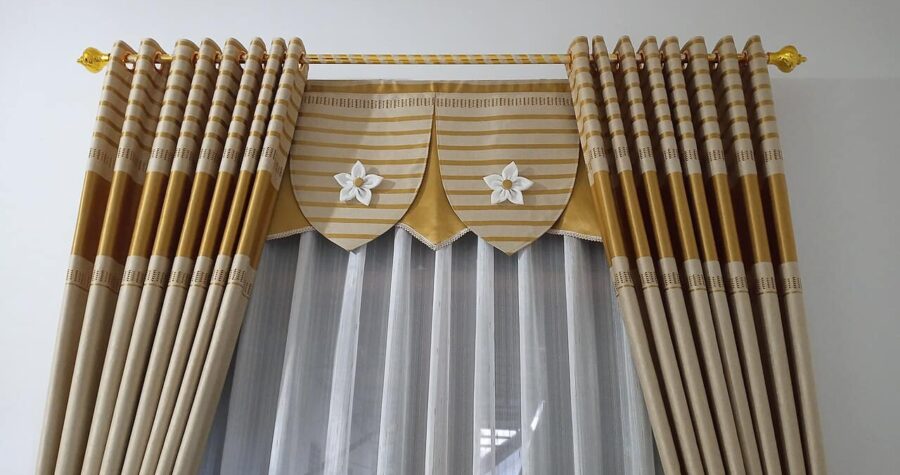 Elegant gold and beige striped curtains with decorative valance, white flower appliqués, sheer underlayer, gold finials on curtain rod, formal drapery treatment for living area