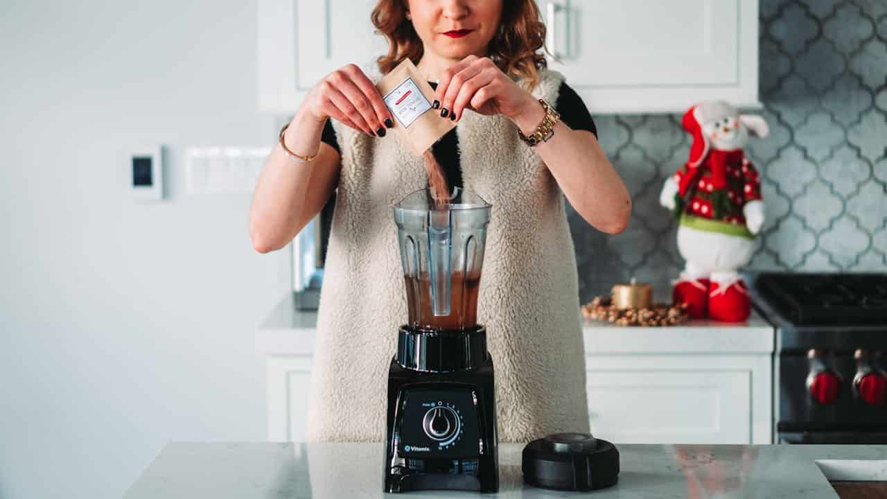 Woman preparing a blended drink, pouring powdered ingredient into a blender, modern kitchen setting, festive snowman decoration in the background, marble countertop, black Vitamix blender