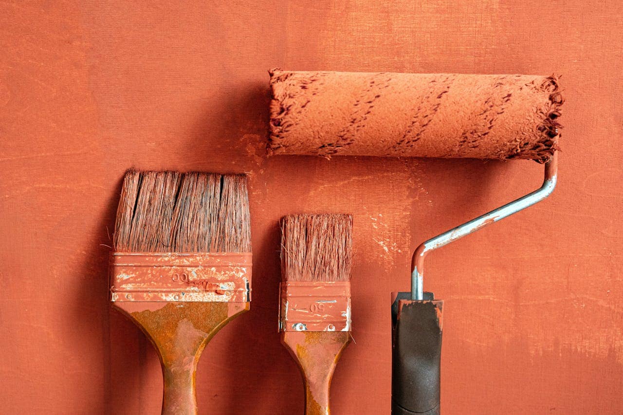 Three paintbrushes and a paint roller covered in bold, rusty orange paint, brushes showing signs of wear, roller handle slightly rusted, textured paint strokes visible on the surface