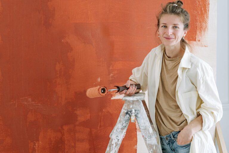 Woman standing on a ladder, holding a paint roller, painting a wall orange, smiling, wearing a beige shirt and white jacket, unfinished paint strokes visible