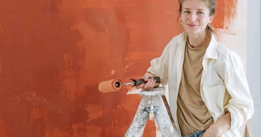 Woman standing on a ladder, holding a paint roller, painting a wall orange, smiling, wearing a beige shirt and white jacket, unfinished paint strokes visible