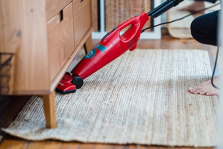 Person vacuuming a beige woven rug, red handheld vacuum cleaner, cleaning under a wooden cabinet, bare foot visible, home interior setting