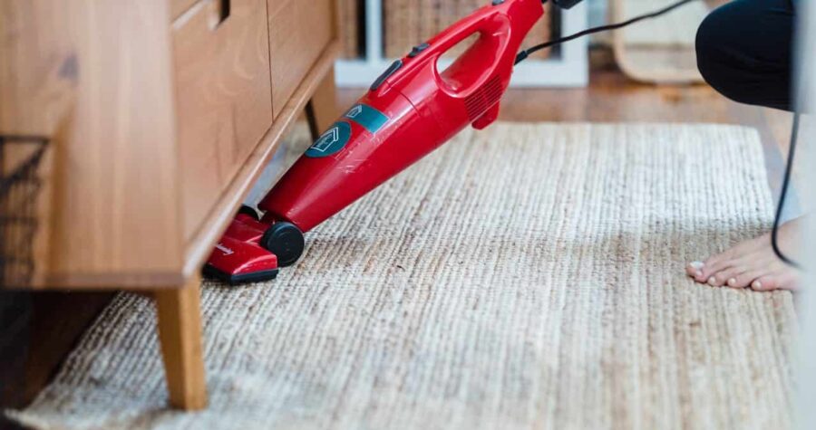 Person vacuuming a beige woven rug, red handheld vacuum cleaner, cleaning under a wooden cabinet, bare foot visible, home interior setting