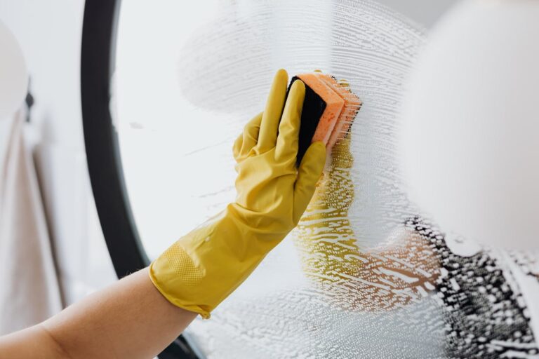 A person wearing yellow rubber gloves scrubbing a soapy mirror with an orange and black sponge, creating streaks of foam on the glass surface