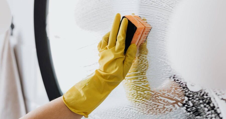 A person wearing yellow rubber gloves scrubbing a soapy mirror with an orange and black sponge, creating streaks of foam on the glass surface