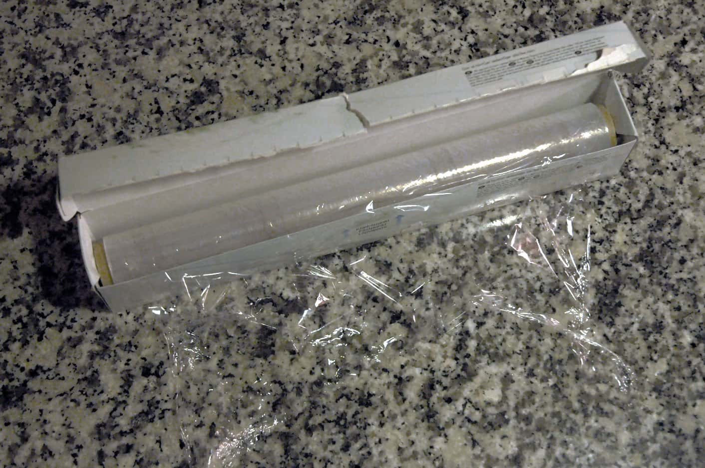 Cling film roll, partially unwrapped, in its box, on a granite countertop, packaging torn slightly, transparent plastic, used for food storage, kitchen item, ready to be used for wrapping or preserving items