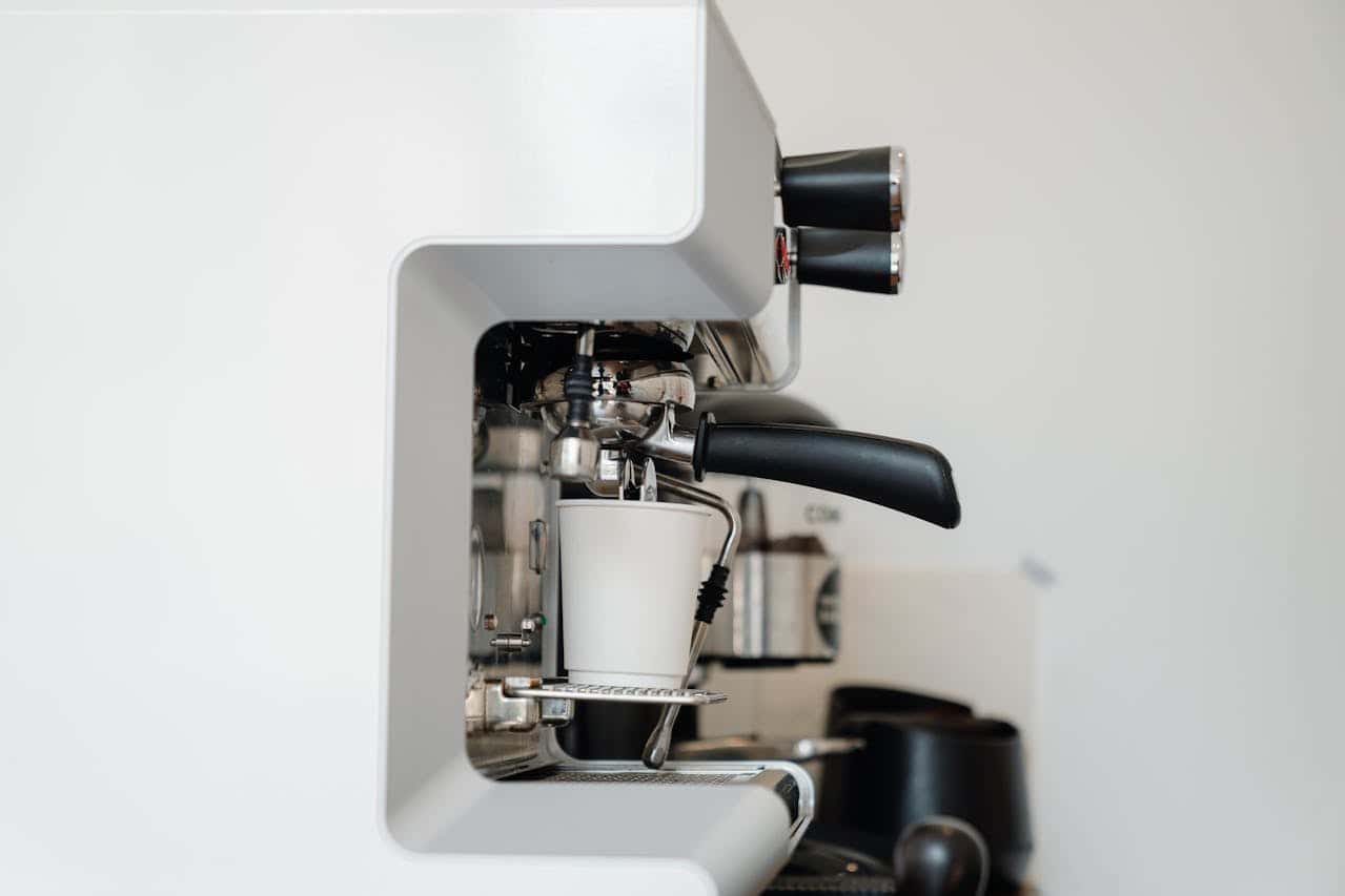 Professional espresso machine, white and sleek design, black portafilter handle, brewing coffee into a white paper cup, stainless steel components, steam wand visible, coffee-making setup, modern kitchen or café setting, background with coffee accessories