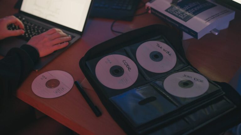 Laptop on a desk, person typing, open CD case with labeled discs, loose CD and pen nearby, software engineering book, dim lighting