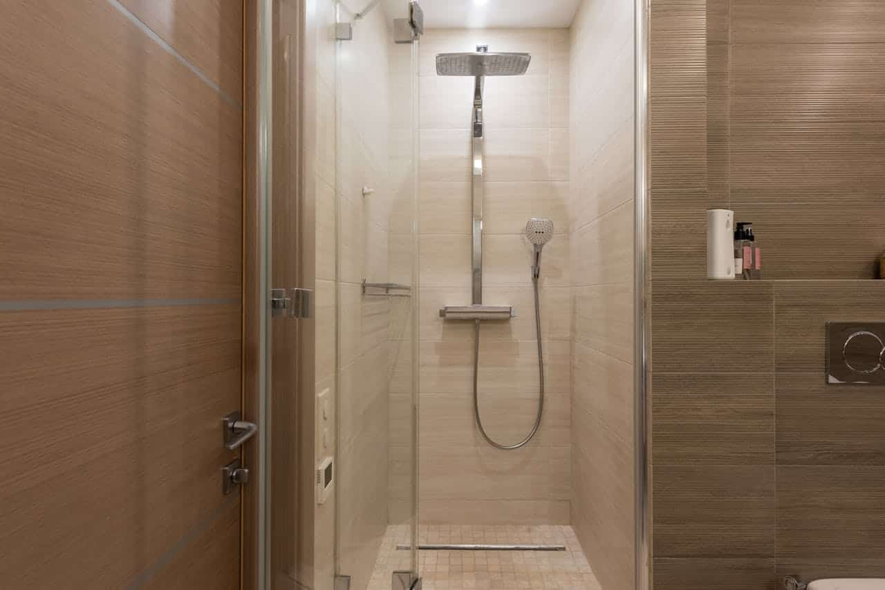 Modern walk-in shower with glass door, beige tile walls, rainfall showerhead, handheld sprayer, linear drain, wooden door entrance, and small storage niche with bath products