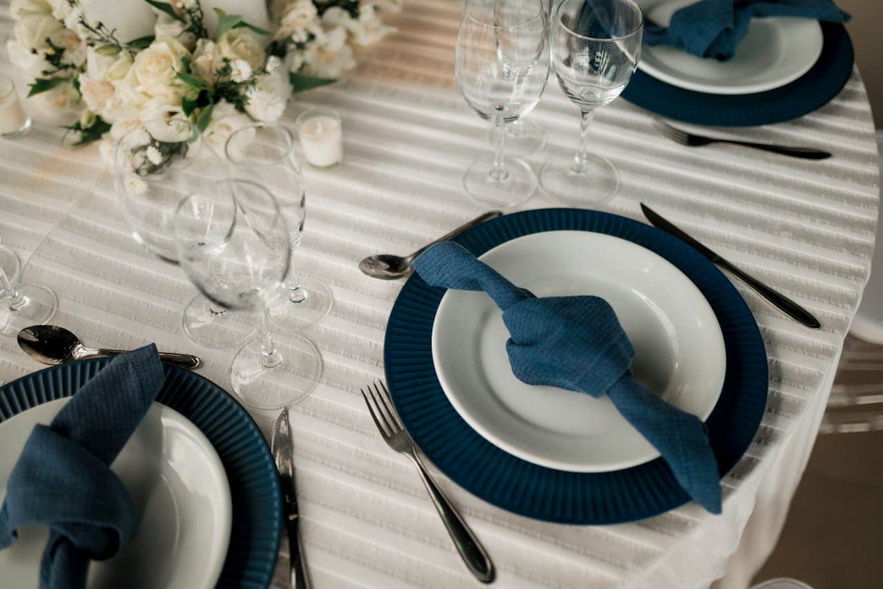 Elegant table setting with white and navy blue color scheme, white plates on blue chargers, folded blue napkins, crystal glassware, silver cutlery, cream-colored floral centerpiece, textured white tablecloth