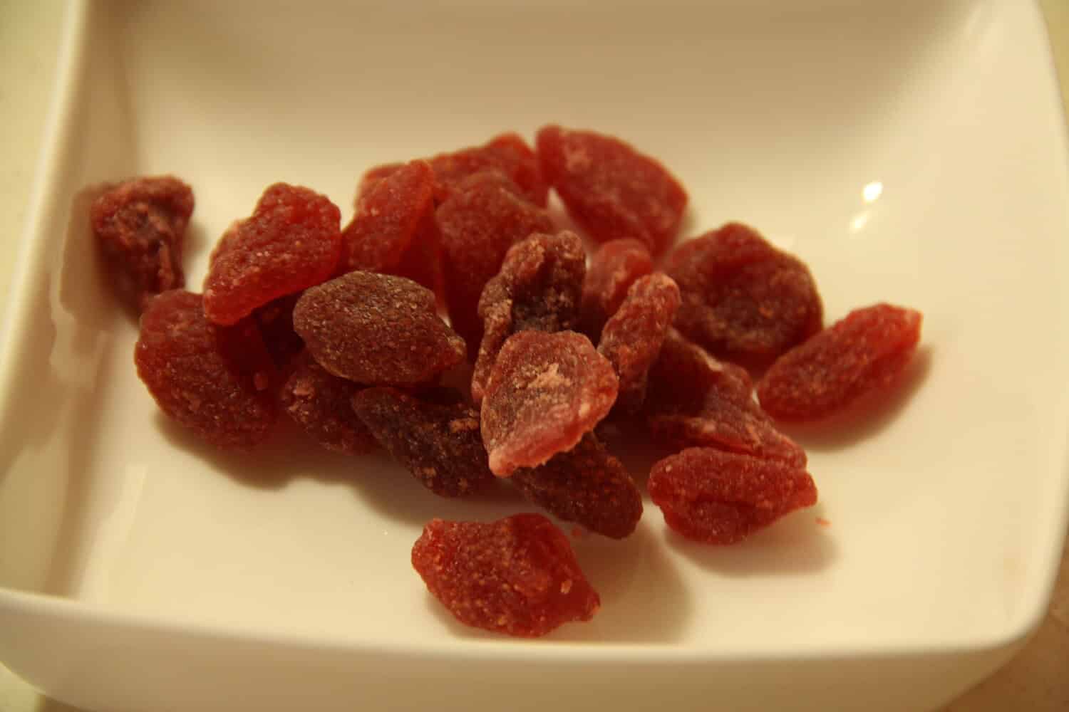A small pile of dried strawberries on a white dish, pieces of dehydrated fruit scattered in various shapes, light texture and color visible
