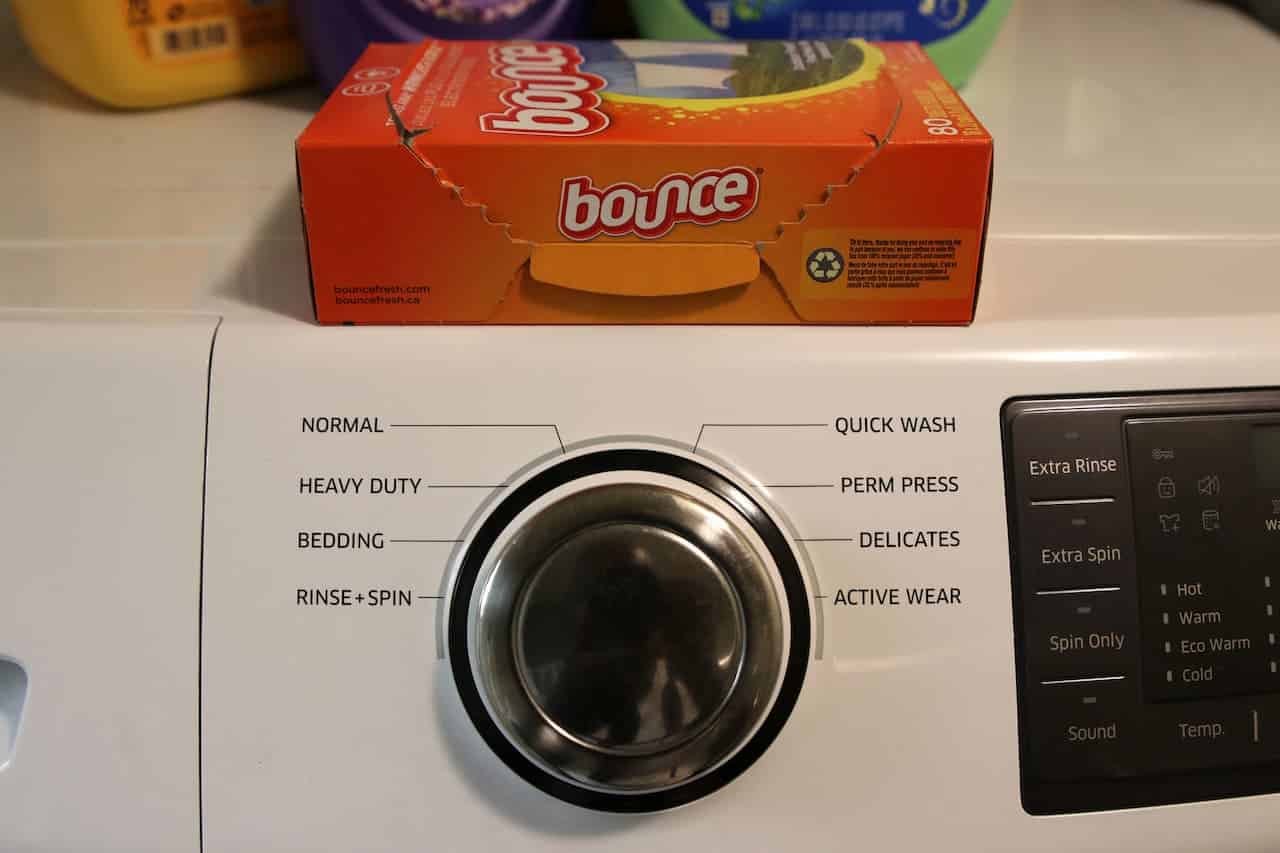 Laundry machine control panel, washing cycle selector knob with various settings, Bounce fabric softener box on top, detergent bottles in the background, clean laundry setup