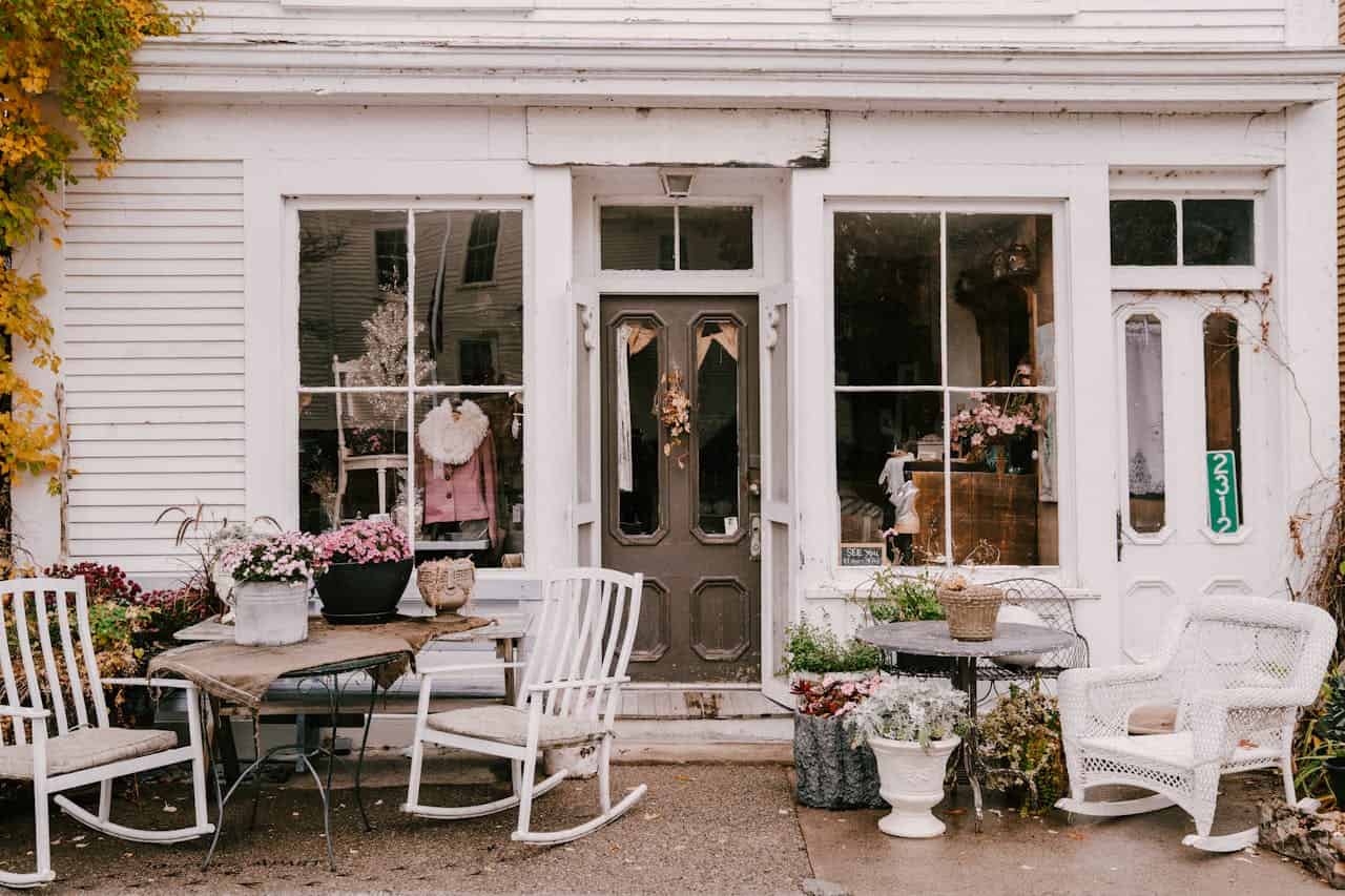 Vintage storefront with white wooden exterior, rustic door with decorative details, large display windows with antique decor, outdoor seating with white rocking chairs