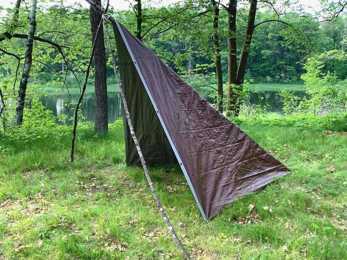 Tent tarp setup, forest camping setup, brown tarp forming a triangle, poles holding tarp up, natural outdoor scenery, trees and water in the background, camping shelter, tarp secured with rope