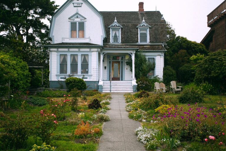 Historic white Victorian house, lush garden with colorful flowers, concrete pathway leading to entrance, wooden chairs on the lawn, overcast sky, greenery surrounding the house, peaceful outdoor setting