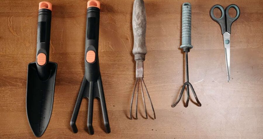 Gardening tools on a wooden surface, includes a trowel, hand rake, cultivator, weeder, and pruning scissors, neatly arranged in a row