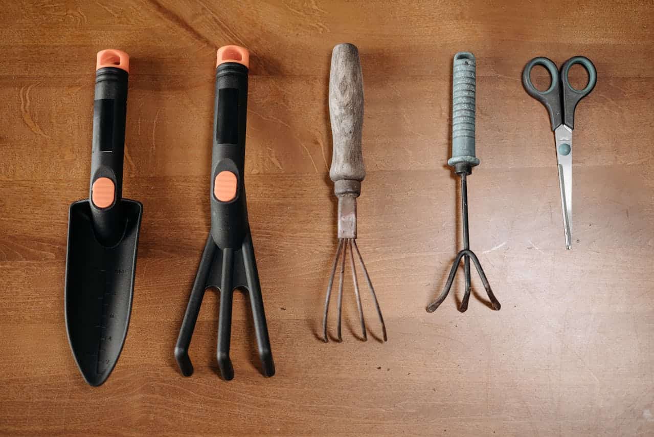 Gardening tools on a wooden surface, includes a trowel, hand rake, cultivator, weeder, and pruning scissors, neatly arranged in a row