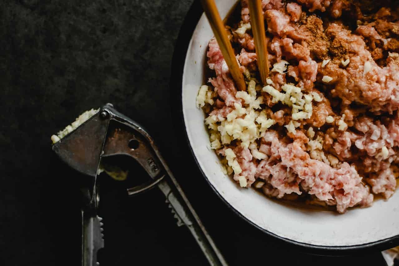 Bowl of raw ground meat, minced garlic, and spices, chopsticks mixing ingredients, garlic press with crushed garlic on a dark surface, food preparation scene