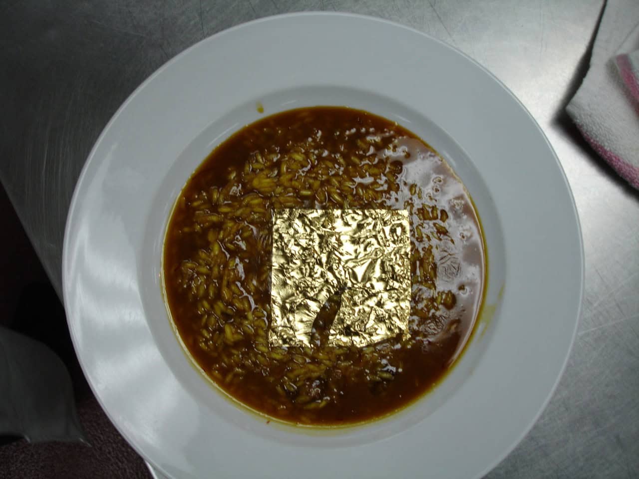 A plate of rice with a rich, brown sauce is topped with a square of edible gold leaf, showcasing a luxurious touch to the dish, the plate is white with a clean design, the setting is likely in a professional kitchen or restaurant, emphasizing gourmet presentation