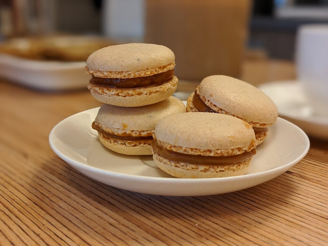 Four caramel macarons on white plate, beige cookie shells with brown filling, wooden table surface, café setting, close-up view
